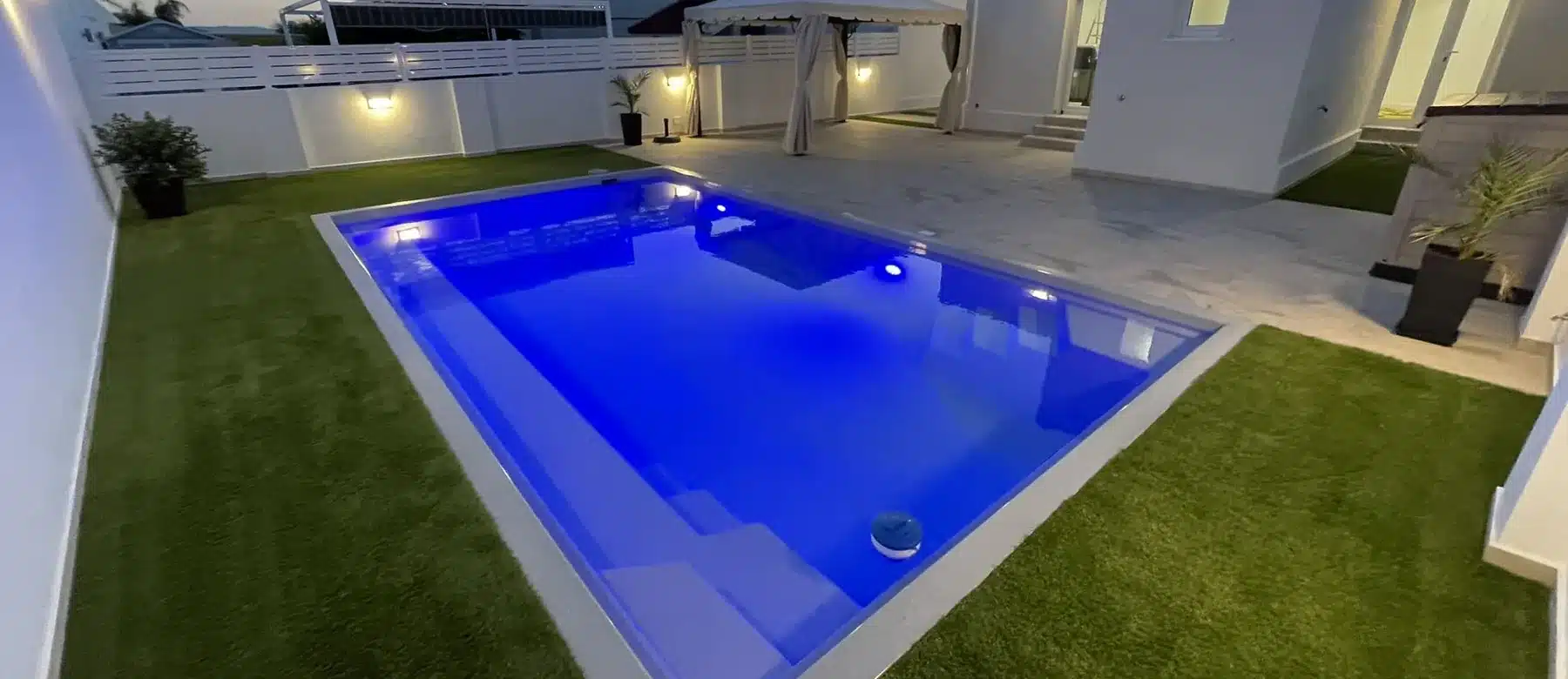 Piscine coque Atlantis 7 éclairée le long d'une maison, en fin de journée