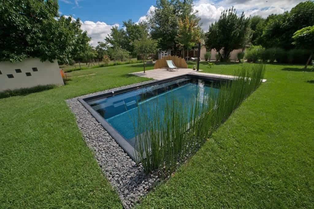 Piscine coque rectangulaire bordée de galets et d'une terrasse en bois, dans un jardin arboré
